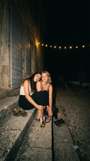 Deux amies assises sur les marches dans la nuit, chaussures enlevées, une bouteille entre elles.