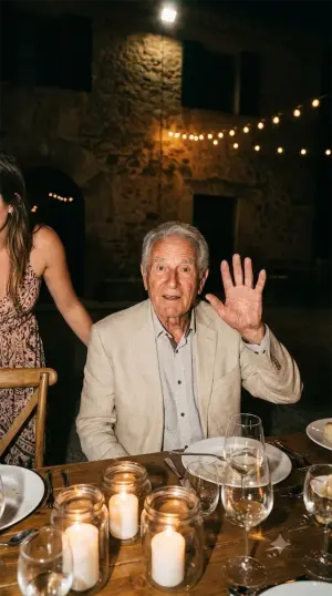 Un grand-père assis à table fait un signe de la main à l’objectif, entre les bougies.